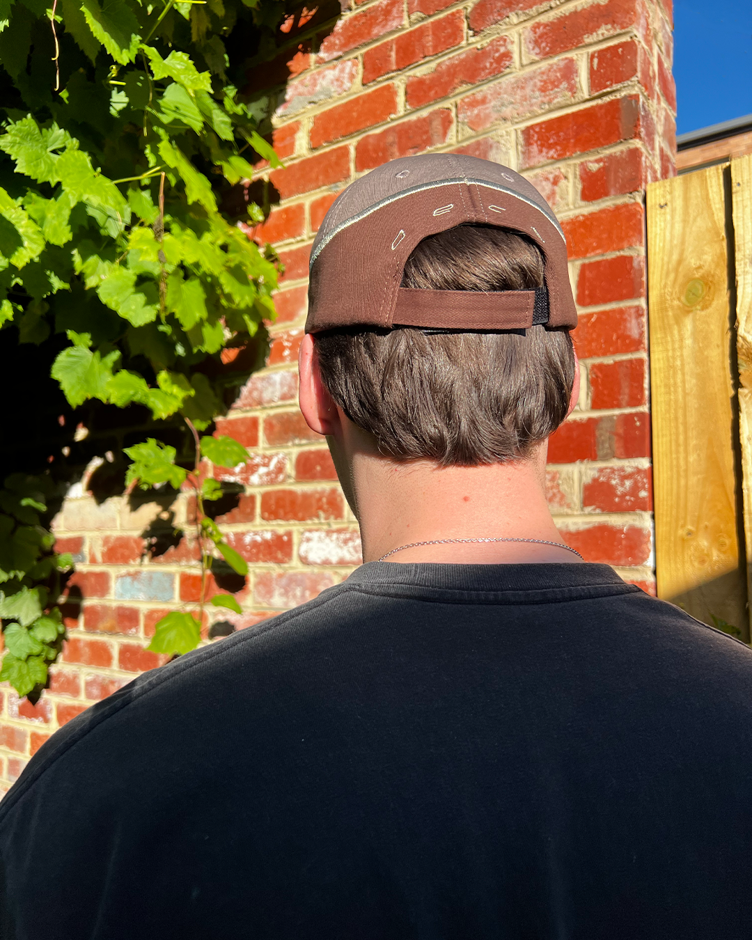 Person wearing a brown cap with a brick wall and greenery in the background