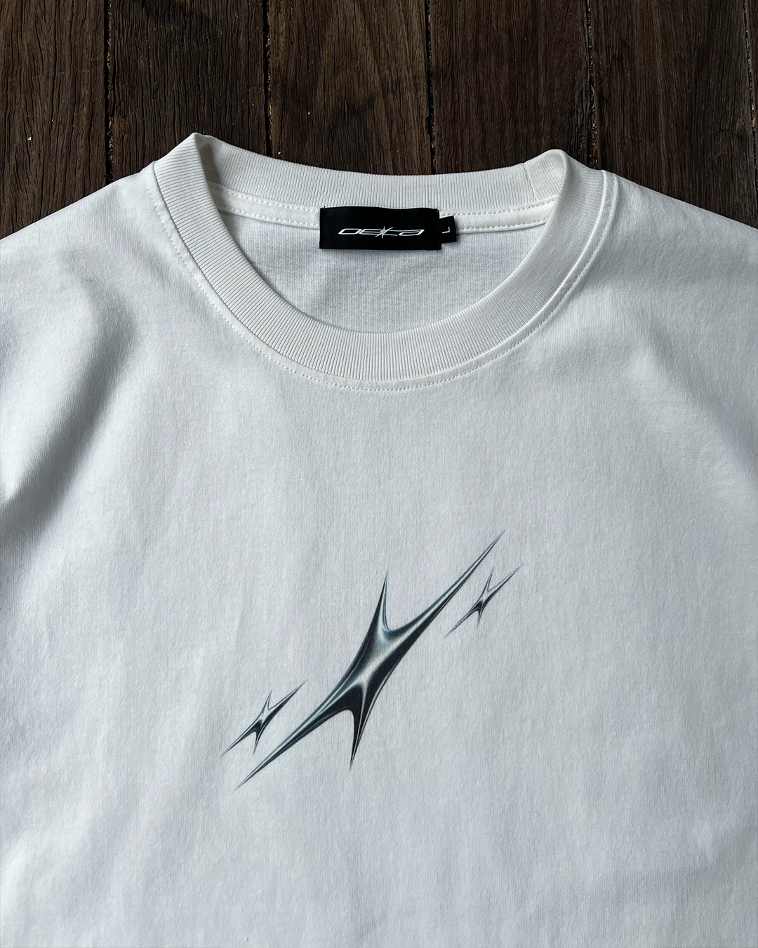 White t-shirt with 3 silver abstract stars design and brand label on a wooden surface